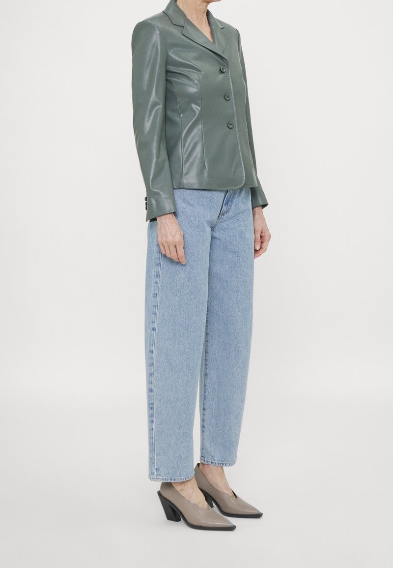 Femme portant un blazer vert brillant boutonné, un jean droit taille haute bleu clair et des talons blocs beiges à bouts pointus.