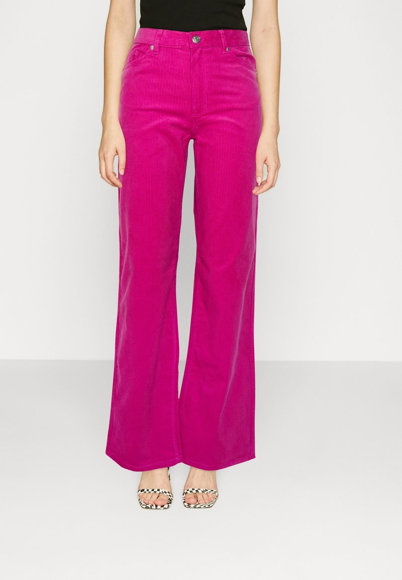 Pantalon large en velours côtelé fuchsia taille haute, avec poches avant et fermeture à bouton, associé à des sandales compensées noires.