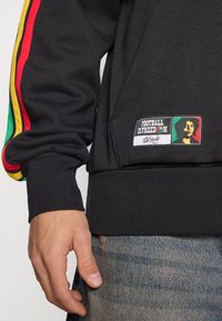 Sweat à capuche noir avec des bandes colorées rouges, jaunes et vertes sur les manches. Il présente un écusson avec l'inscription "Football Is Freedom" et une image de Bob Marley.