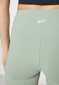 Leggings a vita alta in tessuto verde chiaro con texture liscia; presenta un piccolo logo Nike bianco sul retro. I dettagli delle cuciture sono visibili.