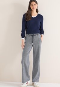 Pull en tricot bleu marine à col en V et pantalon gris à jambes larges avec cordon de serrage, présentant un motif texturé. Des baskets blanches complètent la tenue.