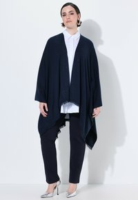 Marineblaue offene Strickjacke aus weichem Stoff mit Fransen; getragen über einem weißen Hemd mit Kragen und dunklen Hosen, ergänzt durch silberne Absätze.