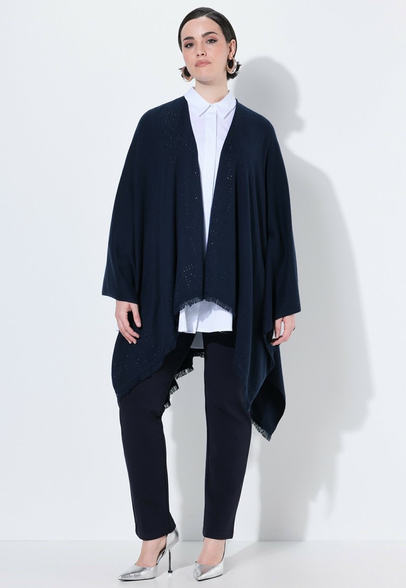 Granatowy kardigan z otwartym przodem, wykonany z miękkiego materiału, z frędzlami; noszony na białej koszuli z kołnierzykiem i ciemnych spodniach, uzupełniony srebrnymi szpilkami.