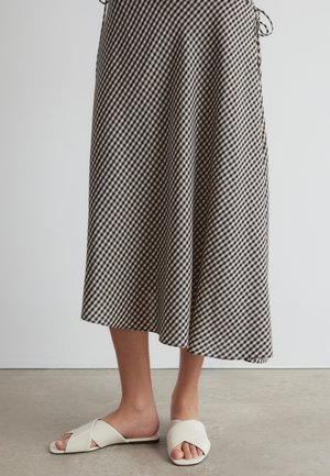 Vue sur la longueur des jambes d'une personne portant une jupe midi à carreaux noir et blanc avec des sandales blanches à enfiler avec des brides croisées.