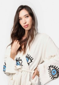 Biely kapucňový plášť s čiernymi a modrými očnými vzormi na rukávoch, vyrobený z mäkkej tkaniny s strapcovými okrajmi a opaskom na viazanie.