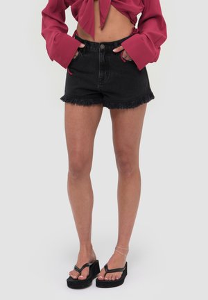Kobieta ubrana w związany czerwony crop top z długim rękawem, czarne poszarpane szorty jeansowe i czarne platformowe klapki, stoi z rękami w kieszeniach.