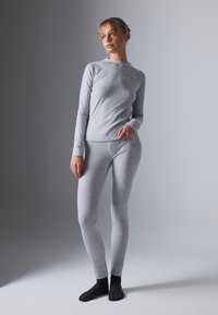 Una donna indossa un top a maniche lunghe grigio chiaro e leggings abbinati con calzini neri, in piedi davanti a uno sfondo grigio semplice.
