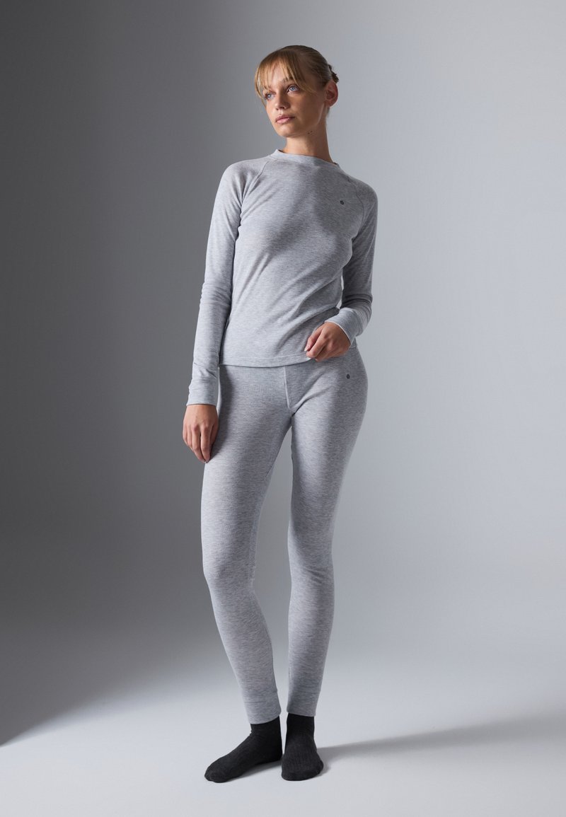 Una donna indossa un top a maniche lunghe grigio chiaro e leggings abbinati con calzini neri, in piedi davanti a uno sfondo grigio semplice.