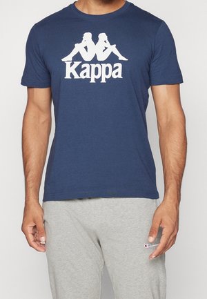 Homme portant un t-shirt Kappa bleu marine avec un logo blanc et un pantalon de survêtement Champion gris clair, debout devant un fond uni.