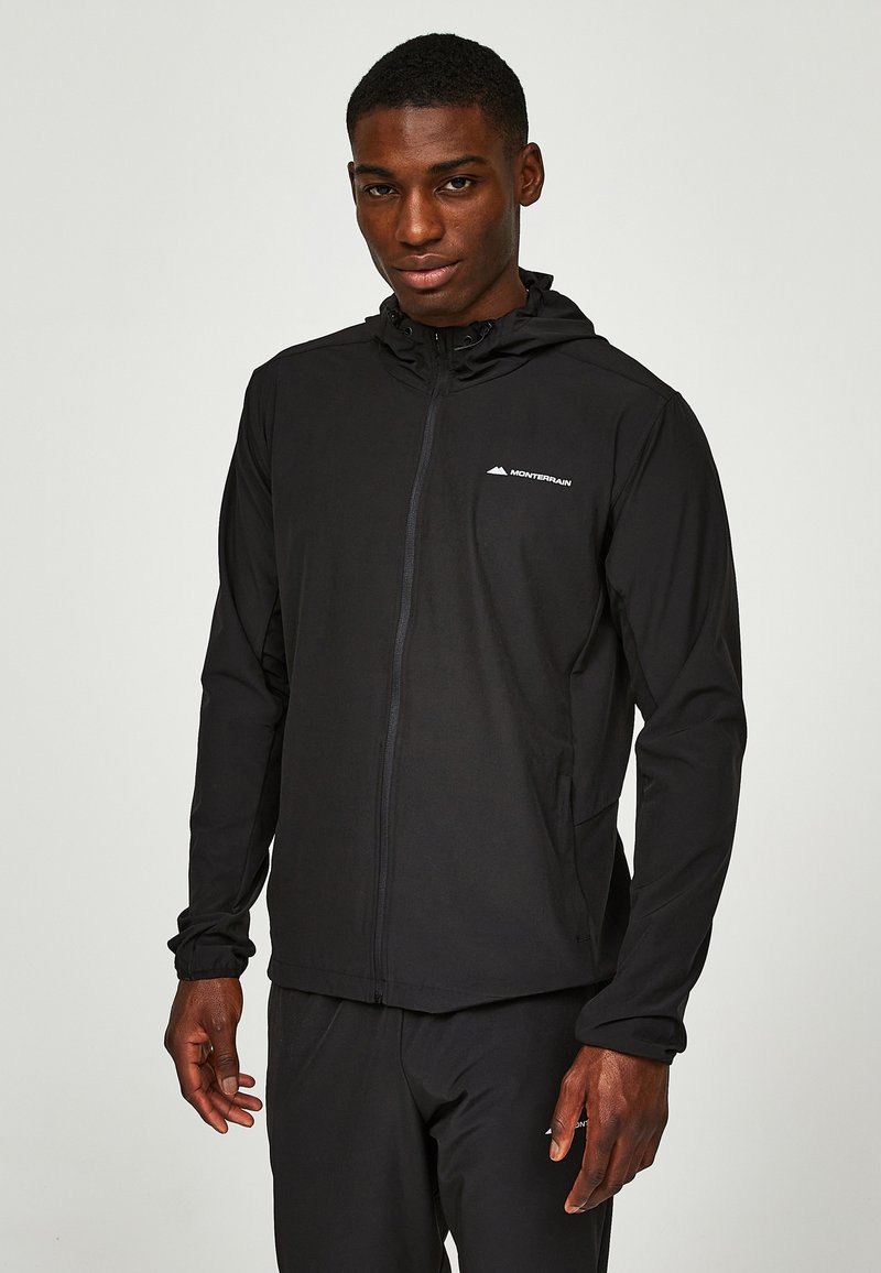Jeune homme portant une veste zippée noire Monterrain avec capuche et un pantalon noir assorti, debout devant un fond uni.