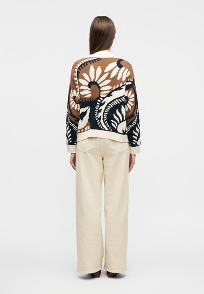 Γυναίκα με μακριά μαλλιά που φοράει καφέ, λευκό και μαύρο πουλόβερ με λουλουδάτο σχέδιο και εκρού παντελόνι με φαρδιά μπάσα, στέκεται γυρισμένη προς τα πίσω.