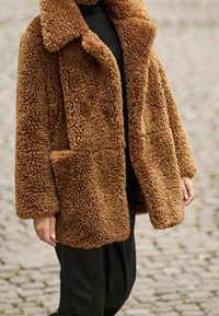 Abrigo marrón de pelo sintético desordenado con cuello ancho, ajuste holgado y bolsillos laterales. La superficie texturizada contrasta con un conjunto negro elegante debajo.