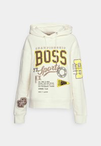 Sweat à capuche crème avec cordon de serrage, présentant des motifs mixtes en jaune et marron, comprenant des slogans et des logos sur le devant et les manches.