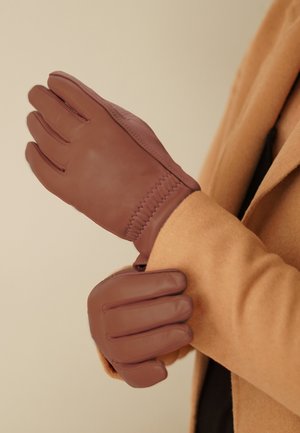 Gants en cuir marron avec une texture lisse, présentant des accents cousus et un design ajusté au poignet, portés sur un manteau marron clair.