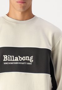 Sudadera gris claro con una franja horizontal gris oscuro. Texto "Billabong" y "Desde mil novecientos setenta y tres" bordado en blanco. Cuello acanalado.