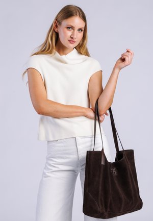 Mujer rubia con una camiseta blanca sin mangas y pantalones blancos sosteniendo un bolso grande de gamuza marrón oscuro con asas para el hombro.