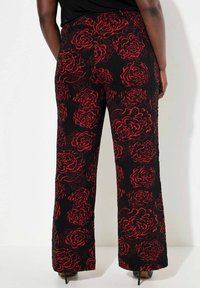 Zwarte broek van een textuurstof, met een rood rozenpatroon. Het ontwerp heeft een wijde pijp en naaddetails aan de achterkant.