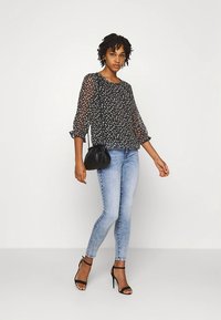 Blusa preta transparente com estampa floral, mangas três quartos. Jeans skinny azul claro. Modelo com bolsa preta e saltos com tiras.