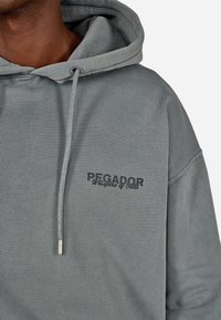 Pelēkas kapuces džemperis ar melnu priekšējo logotipu, aukliņu aizdari un mīkstu, struktūru audumu ar brīvu piegulšanu.