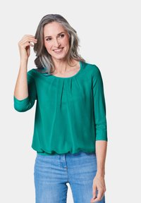 Groene, lichte blouse met 3/4 mouwen, geplooide details bij de halslijn en een relaxte pasvorm, gecombineerd met lichtblauwe jeans.