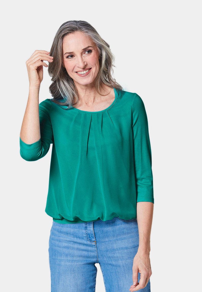 Groene, lichte blouse met 3/4 mouwen, geplooide details bij de halslijn en een relaxte pasvorm, gecombineerd met lichtblauwe jeans.