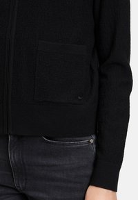 Zwarte zip-up jas van gestructureerde stof, met een voorzak en geribbelde manchetten. Gecombineerd met donkerdenim jeans.