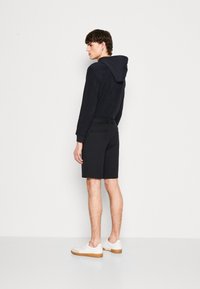 Marinblå hettegenser med teksturert overflate, kombinert med svarte shorts i glatt stoff med baklommer. Hvite sneakers kompletterer antrekket.