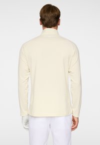 Homme portant une chemise à manches longues crème et un pantalon blanc, vu de dos sur un fond clair uni.