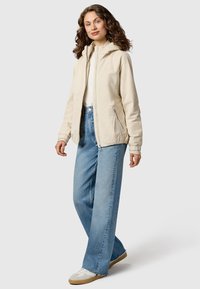 Vrouw met krullend bruin haar, gekleed in een beige hooded jas, wit shirt, lichtblauwe wijde jeans en witte sneakers, staand tegen een witte achtergrond.