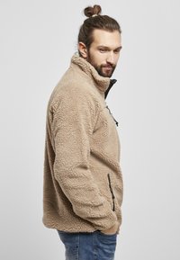 Beige fleece jakke med høj krave, lynlåslommer foran og sidelommer. Struktureret stof og afslappet pasform.