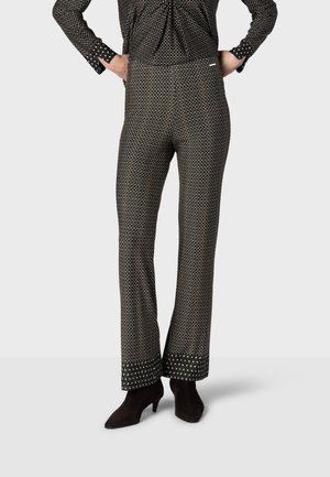 Femme portant un pantalon noir évasé à petits carrés blancs et des bottines noires à talons, mains posées sur les hanches.