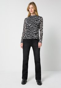 Langärmliges Oberteil mit einem schwarz-grauen Zebra-Muster, aus leichtem, durchscheinendem Stoff gefertigt. Kombiniert mit engen schwarzen Denim-Jeans und schwarzen Schuhen.