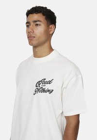 Biała bawełniana koszulka z okrągłym dekoltem, z czarnym napisem graficznym "Good for Nothing" na lewej stronie klatki piersiowej.