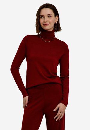 Kvinde iført en tætsiddende vinrød turtleneck med lange ærmer og matchende bukser, smilende og poserende med den ene hånd i lommen.