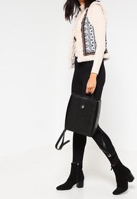 Svart läderryggsäck med texturerad yta, fyrkantig form, bärhandtag och justerbara remmar. Modellen bär en krämfärgad tröja och svarta byxor.