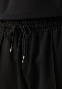 Svarta joggingsbyxor med elastisk midja, som har en dragsko med silverfärgad ändar och sidofickor. Mjuk tygtextur synlig.