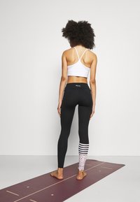 Černé legíny s kontrastním leopardím potiskem a pruhovaným vzorem na levé noze, spárované s bílou sportovní podprsenkou. Naboso na cvičební podložce.