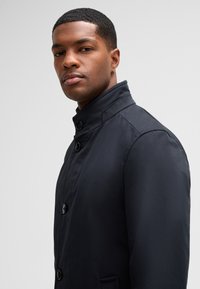 Navy blue coat with a high collar, featuring two front buttons and side pockets. Smooth fabric texture with a tailored fit.