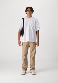 White cotton t-shirt with a small black logo, beige relaxed-fit pants, and cream sneakers with red accents; black crossbody bag.