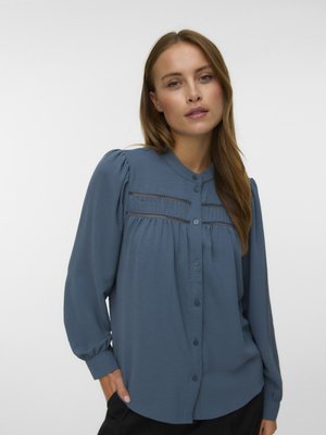 Nő hosszú ujjú, gombos és csipkedíszes kék blúzban, világos szürke, egyszerű háttér előtt áll.