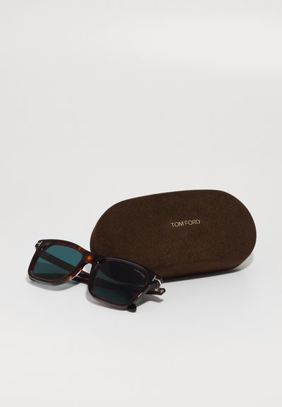 Lunettes de soleil rectangulaires en écaille avec verres foncés à côté d’un étui Tom Ford marron avec inscription dorée sur une surface blanche.