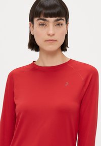Červený top s dlhým rukávom vyrobený z hladkej látky; má okrúhly výstrih a jemné logo na hrudi; prešívanie na ramenách.