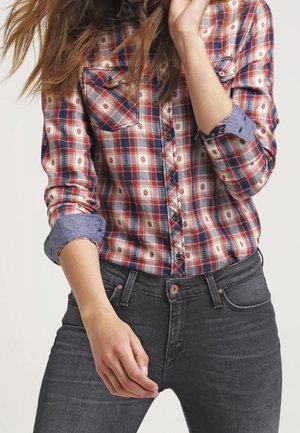 Vrouw draagt een rood-blauw geruit overhemd met opgerolde mouwen en zwarte jeans, poseert met één hand bij haar hoofd.