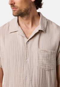 Camicia a maniche corte color beige chiaro con bottoni, realizzata in tessuto testurizzato. Presenta una tasca a toppa con ricamo "SEASIDE" e bottoni bianchi.