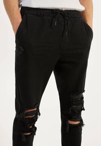 Mężczyzna ubrany w czarne zniszczone dżinsy z wieloma dziurami oraz mający ręce w kieszeniach, w zestawieniu z białą koszulką.