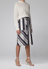 Weißer gestrickter Pullover mit Rundhalsausschnitt, kombiniert mit einem gestreiften Rock in Marineblau und Weiß, der eine Seitentasche und einen gebundenen Taillenbund hat, ergänzt durch helle Absätze.