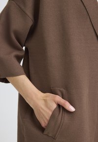 Ruda mezginė apranga su trimis ketvirtadaliais rankovėmis, turinti šoninį kišenę. Tekstūra yra lygi ir šiek tiek struktūruota.