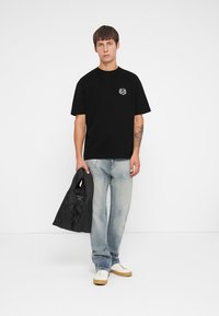 Zwarte T-shirt met korte mouwen en een ronde grafiek op de borst, gecombineerd met lichtblauwe jeans en een zwarte puffer tas. Witte sneakers met beige zolen.