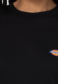 Zbliżenie na osobę w czarnej koszulce typu crewneck z małym, kolorowym logo Dickies po prawej stronie klatki piersiowej.