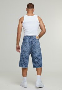 Shorts de mezclilla con un patrón geométrico, color azul y corte recto. Presentan detalles de parches en la cintura y en los bolsillos traseros. Zapatillas blancas.